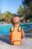A baby is sitting facing a pool, away from the camera, wearing a mango orange Infant Warm Belly Wetsuit, showing off adjustable Velcro shoulder straps and a Velcro diaper flap.