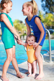 Two little girls are standing in front of a pool looking at each other and laughing while holding a smiling baby. The little girls are wearing Children's Warm Belly Wetsuits and the baby is wearing an Infant Warm Belly Wetsuit. 