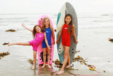 Three smiling little girls are posing on the beach in front of the waves with two boogie boards. All three are wearing Children's Warm Belly Wetsuits in varying colors.
