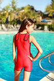 A little girl is facing the pool, head turned looking to the side, with one hand on her hip. She is wearing a red children's custom size Warm Belly Wetsuit.