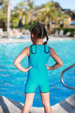 A little girl is standing with her hands on her hips facing a pool, away from the camera, wearing a turquoise children's Warm Belly Wetsuit, showing off the adjustable Velcro shoulder straps.