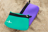 Two Neoprene Beach Pouches laying in the sand on a beach, one turquoise and the other purple. 